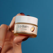 A person holding a small container of Emirates Lip Balm to their lips, suggesting hydration or care for the lips. Xcosmetics | cream x emirates